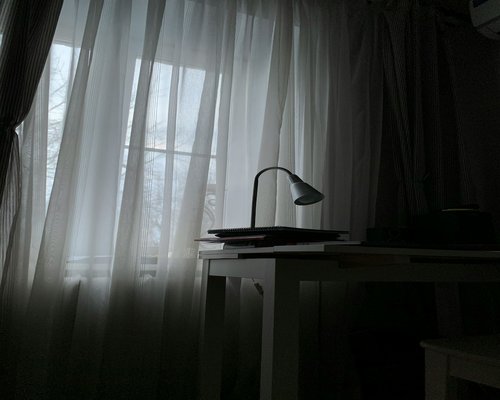 Soft light entering through a curtained window onto a tidy desk with a small desk lamp and papers open for reading